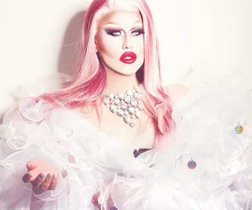 Drag queen in elaborate white gown with pink hair, bold makeup, red lips, and sparkling necklace, posing elegantly.