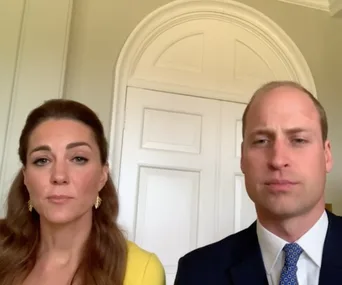 A man in a suit and a woman in a yellow top are sitting in a room with a white door and arched window behind them.