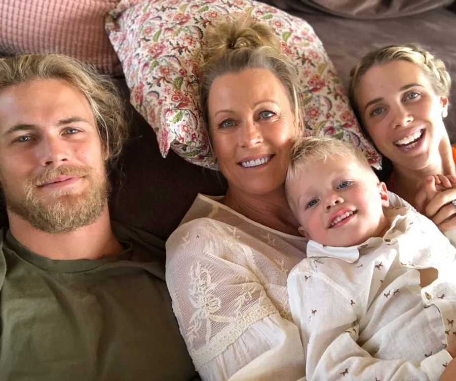 A smiling family with a woman, two young adults, and a child lying on a couch with floral and pink pillows.