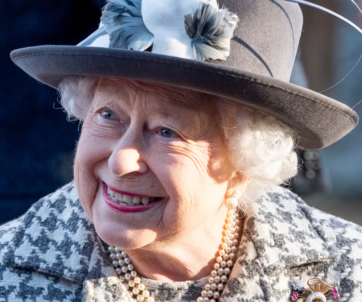 A smiling elderly woman wearing a gray hat and pearl necklace, in a checkered coat.