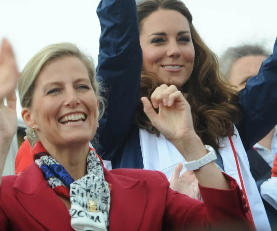 Two women smiling and waving, one wearing a red coat and scarf, the other in a dark jacket.