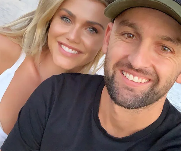 A smiling couple taking a selfie outdoors, with the woman in white and the man in a black shirt and cap.