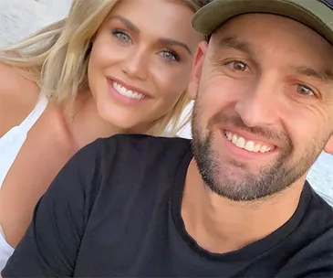 A smiling couple taking a selfie outdoors, with the woman in white and the man in a black shirt and cap.