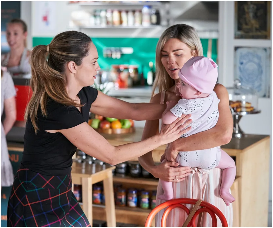 Two women in a tense moment, one holding a baby in a kitchen setting.
