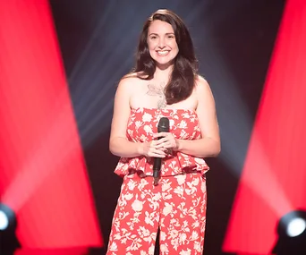 Singer in red floral dress holds a microphone on stage with red spotlights in the background.