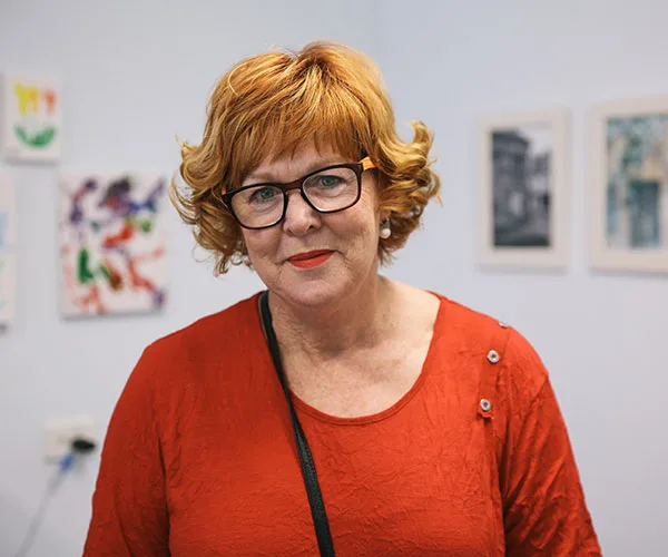Middle-aged woman with red hair and glasses in red top, standing in a room with colorful artwork on the walls.