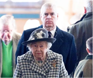 Elderly woman in houndstooth coat and decorative hat, pearls, looking forward; man in suit behind, indoors.
