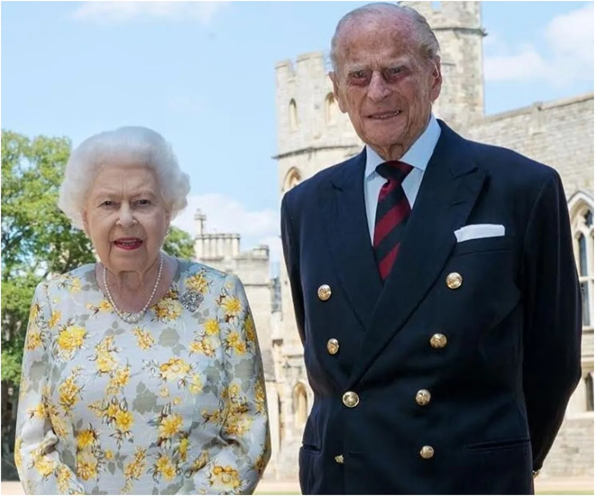 Two elderly individuals dressed formally, standing outside a castle with a tower in the background.