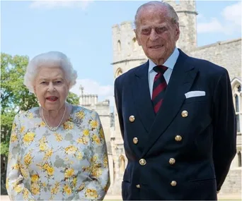 Two elderly individuals dressed formally, standing outside a castle with a tower in the background.
