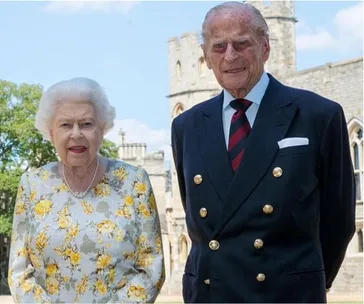 Two elderly individuals dressed formally, standing outside a castle with a tower in the background.
