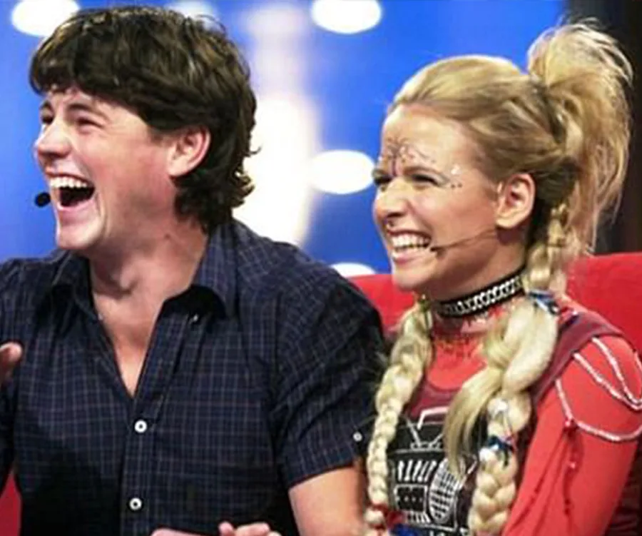 A man and woman with braided hair, both laughing on a brightly lit stage.