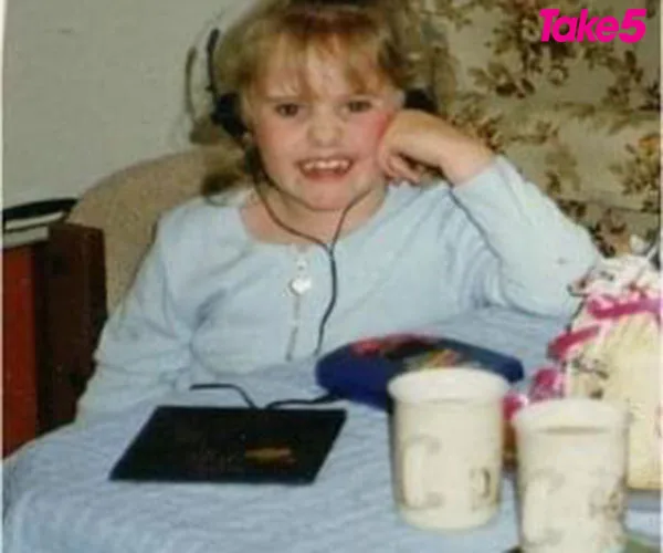 A young girl with headphones smiles while sitting on a couch, with a CD player and two cups on a table in front of her.