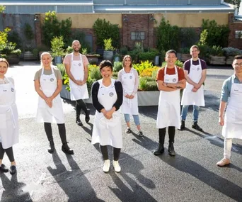 Eight MasterChef 2020 contestants wearing aprons, standing outdoors in a garden setting, smiling for the camera.