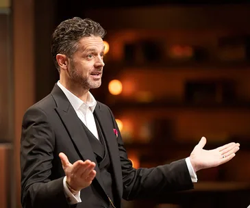 Man in a suit gesturing with open hands in a warmly lit room.