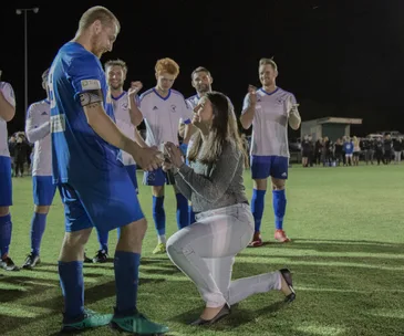 Woman proposes to a man in a soccer uniform on a field, teammates clapping in the background.