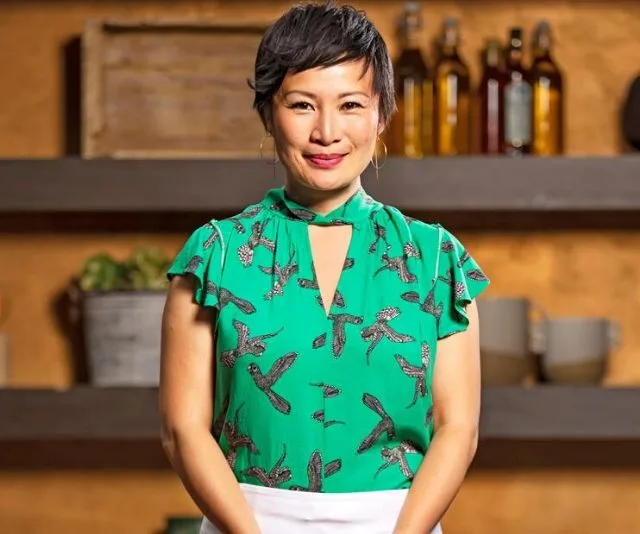 A woman in a green bird-patterned blouse stands smiling in a kitchen setting.