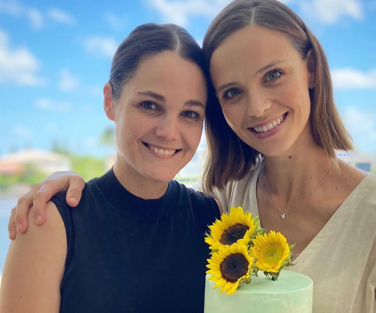 Two women smiling, one holding a cake adorned with sunflowers against a bright blue sky backdrop.