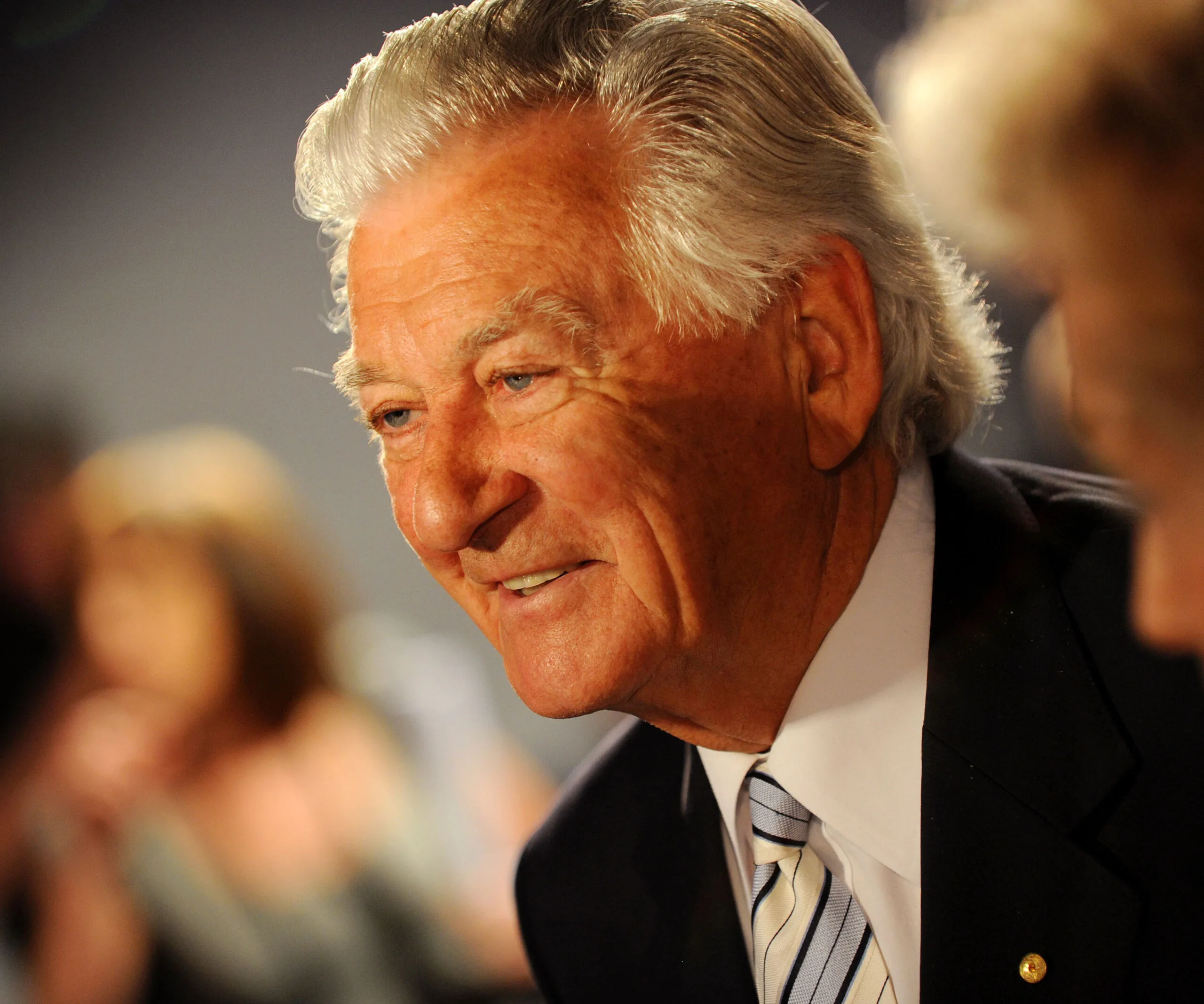 Elderly man with white hair and suit, smiling warmly in a dimly lit setting.