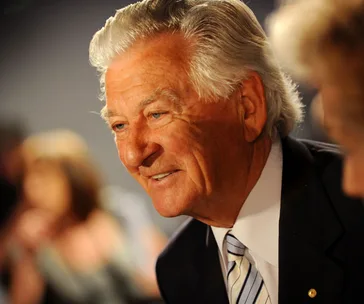 Elderly man with white hair and suit, smiling warmly in a dimly lit setting.