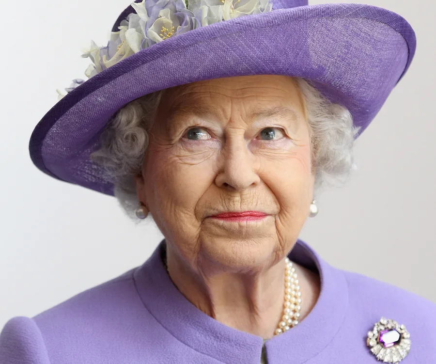 Elderly woman in a purple outfit and hat with flowers, wearing pearl earrings and necklace, glancing upwards.