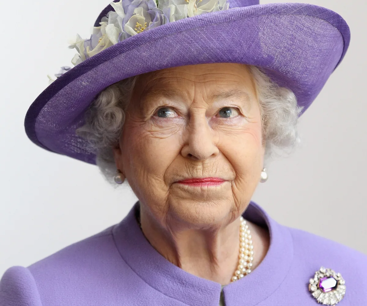 Elderly woman in a purple outfit and hat with flowers, wearing pearl earrings and necklace, glancing upwards.