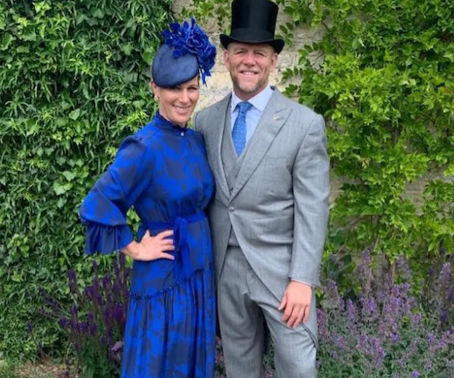 A couple in formal attire, woman in a blue dress and hat, man in a gray suit and top hat, standing outdoors by greenery.