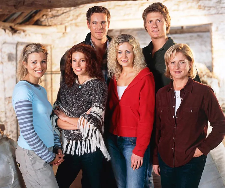 A group photo of six cast members from "McLeod's Daughters" in a rustic setting, smiling and standing close together.