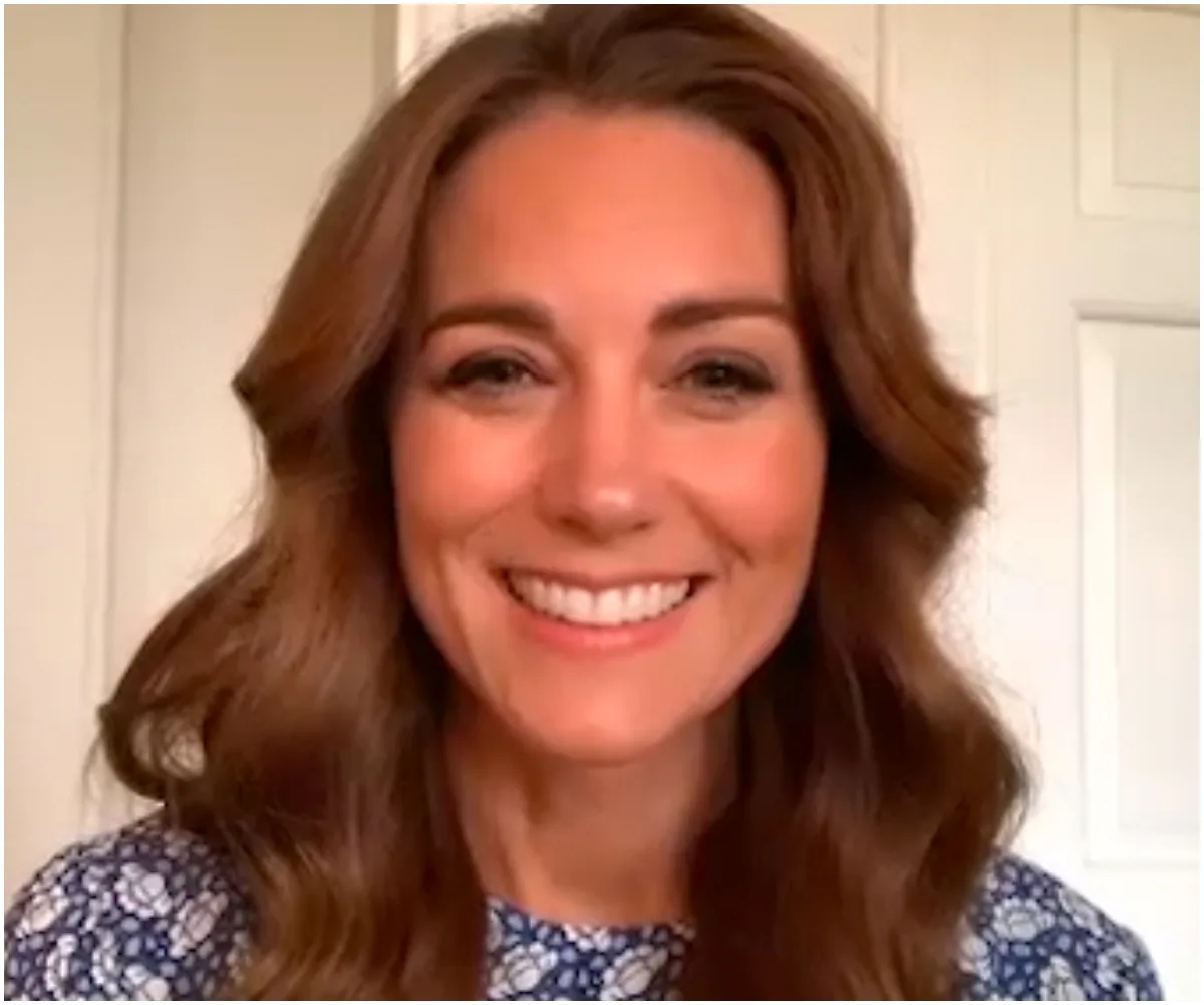 Smiling woman with long brown hair wearing a blue floral top, in front of a light-colored background.
