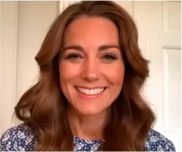 Smiling woman with long brown hair wearing a blue floral top, in front of a light-colored background.