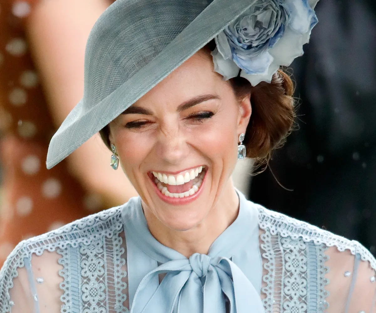 A woman in a blue lace dress and matching hat with a floral design smiles joyfully at an outdoor event.