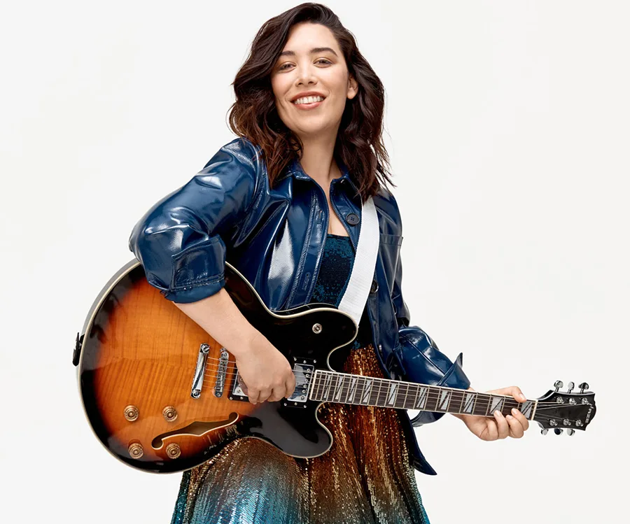 Smiling woman in a blue jacket plays an electric guitar against a plain background.