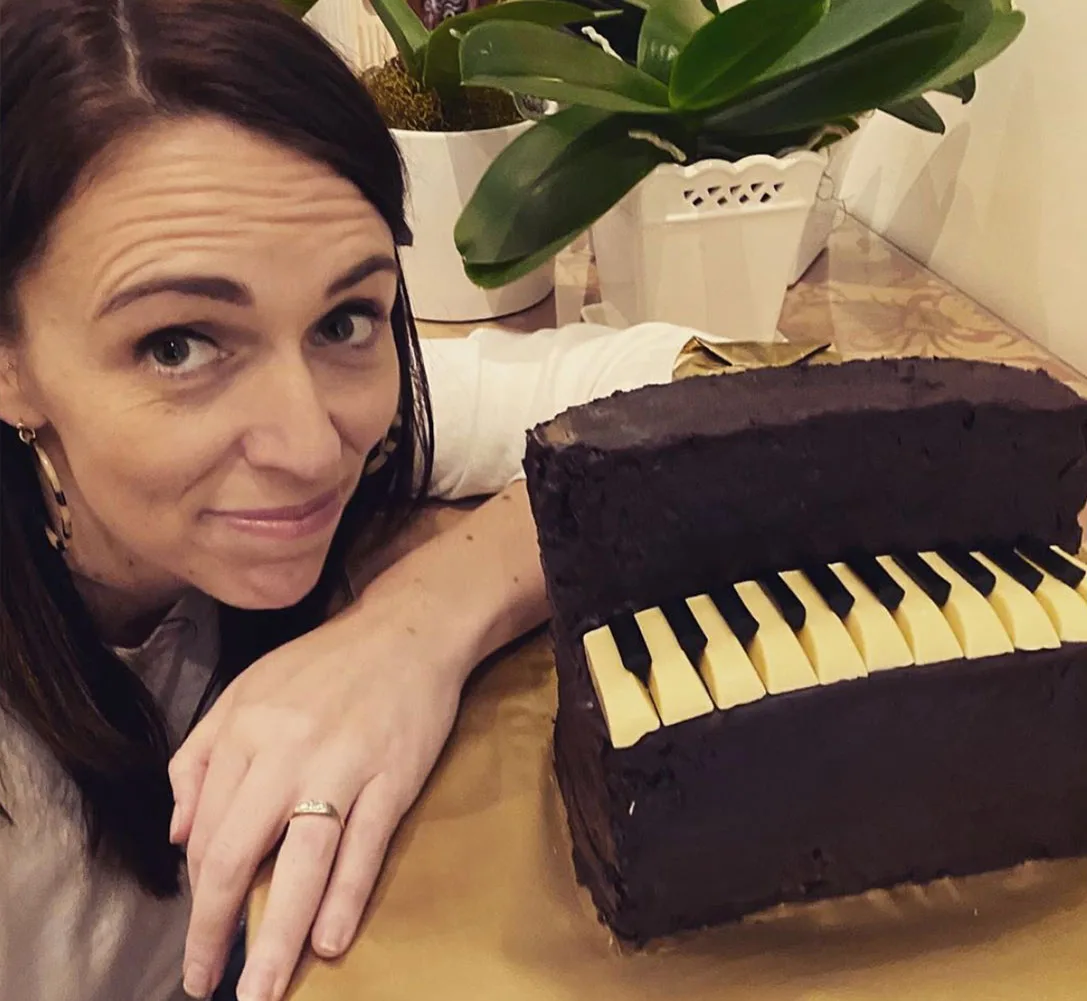 A person smiling next to a cake shaped like a piano with white keys, surrounded by green plants.