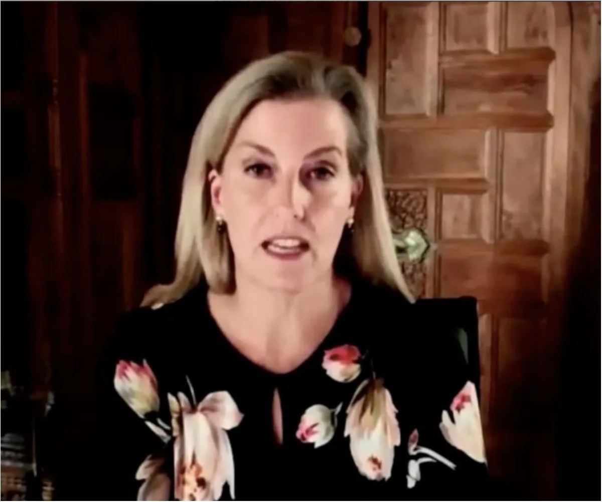 Woman speaking during an online call, wearing a floral blouse, with a wooden background.