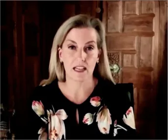 Woman speaking during an online call, wearing a floral blouse, with a wooden background.
