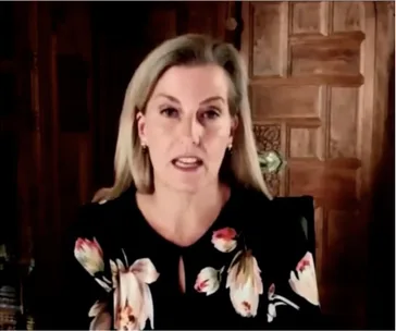 Woman speaking during an online call, wearing a floral blouse, with a wooden background.