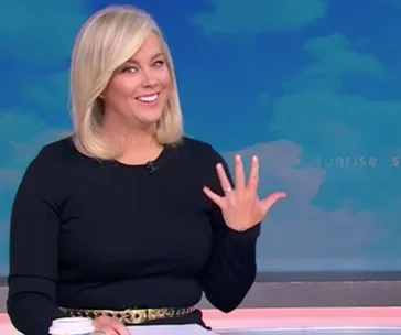 Woman smiling, showing off her engagement ring on a TV set, wearing a black top and holding a coffee cup.