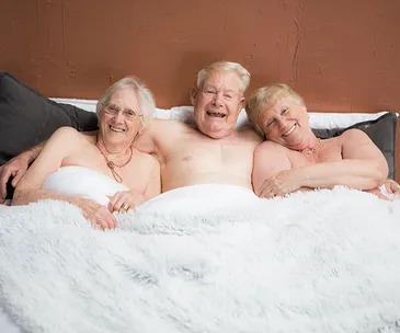 Elderly trio smiling under a blanket, posing together for a calendar.