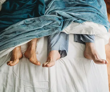 Two pairs of bare feet sticking out from under a blue blanket on a bed.
