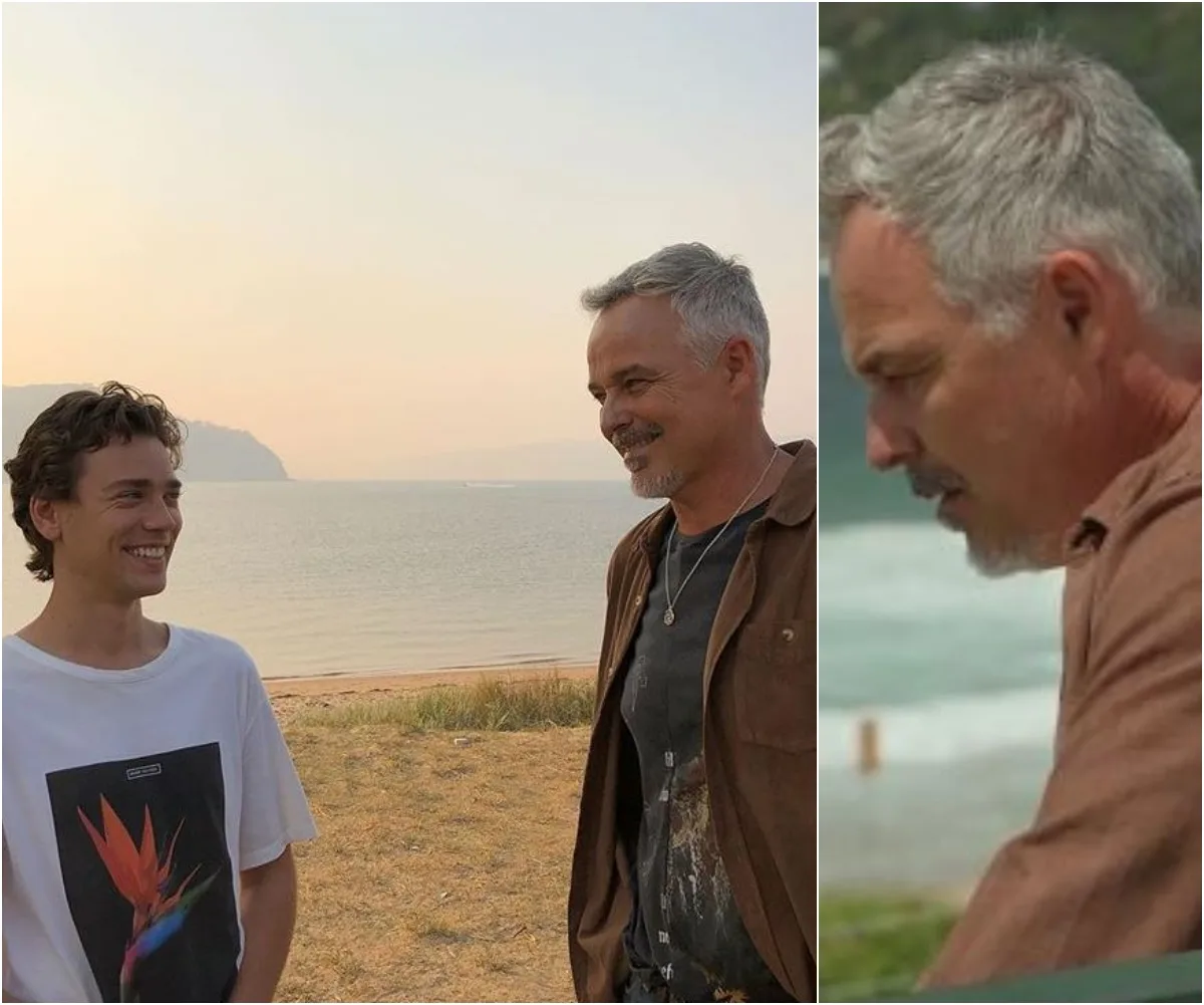 Two people smiling at each other on a beach, with the ocean and hills in the background.