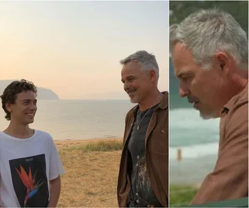 Two people smiling at each other on a beach, with the ocean and hills in the background.