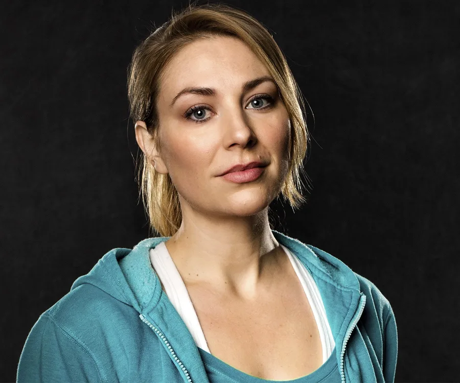 A woman in a teal hoodie with a neutral expression against a dark background.