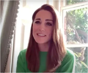 A woman in a green dress smiles during a video call, seated by a window with greenery visible outside.