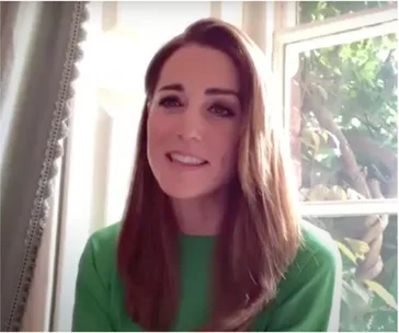 A woman in a green dress smiles during a video call, seated by a window with greenery visible outside.