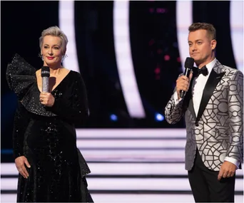 Two hosts onstage with microphones, wearing formal attire: a black dress and a patterned silver suit.