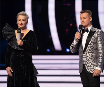 Two hosts onstage with microphones, wearing formal attire: a black dress and a patterned silver suit.