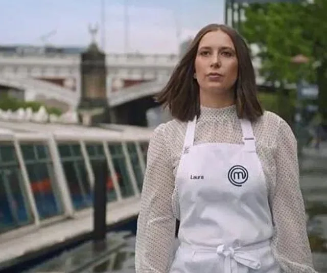 MasterChef contestant in apron outdoors by a riverside with a bridge in the background.