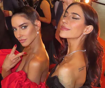 Two women posing stylishly on a red carpet, one with peace sign, both wearing evening gowns and makeup.