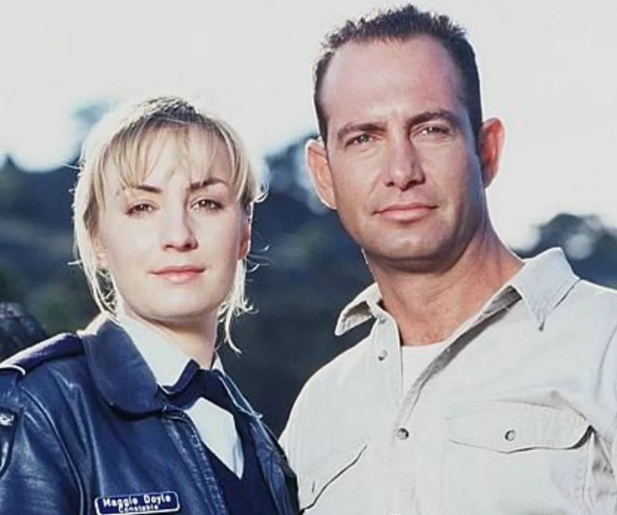 Maggie Doyle and a male character from "Blue Heelers" TV show standing outdoors, both looking at the camera.