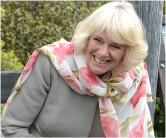 Duchess Camilla smiling, wearing a floral scarf and grey outfit, sitting outdoors.