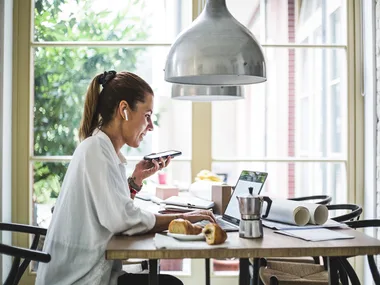 A woman working from home at a laptop, holding a phone, with earphones in, seated at a table with a croissant and papers.